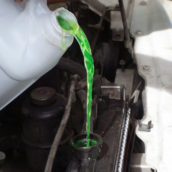 Bright green coolant is poured from a white plastic jug into a car radiator opening under the hood during maintenance.