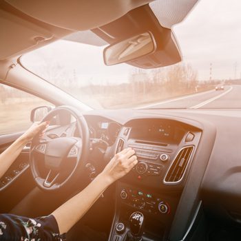 Driver’s hands on steering wheel and radio controls inside a modern car traveling down a sunlit, mostly empty highway.