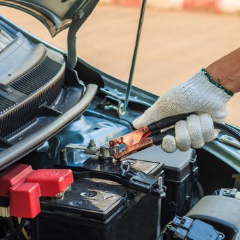 Mechanic wearing a white glove attaches a jumper cable clamp to a car battery terminal under the open hood safely.