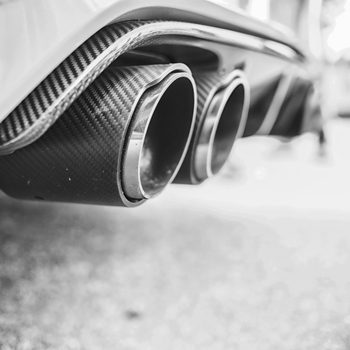 Black and white close-up of dual chrome car exhaust pipes extending from a carbon fiber bumper above pavement.