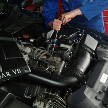 Mechanic in blue coveralls working on a Jaguar V8 engine bay, using a screwdriver to adjust components under hood.