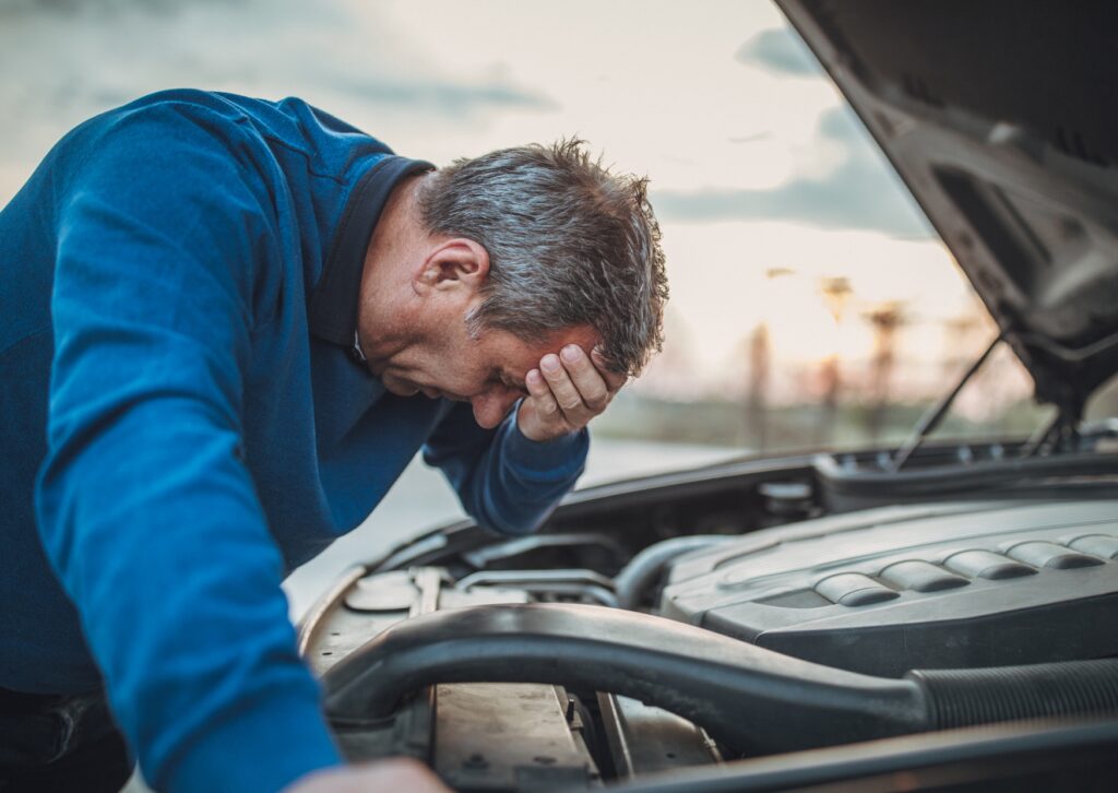 Worried driver leans over open hood with hand on forehead, facing breakdown from fuel system issues in engine bay.