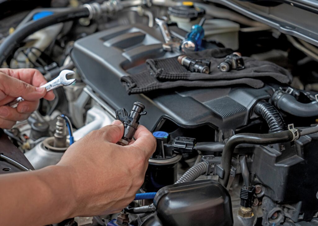 Mechanic holds fuel injector and wrench over open engine bay, with tools and parts laid on engine cover for repairs.