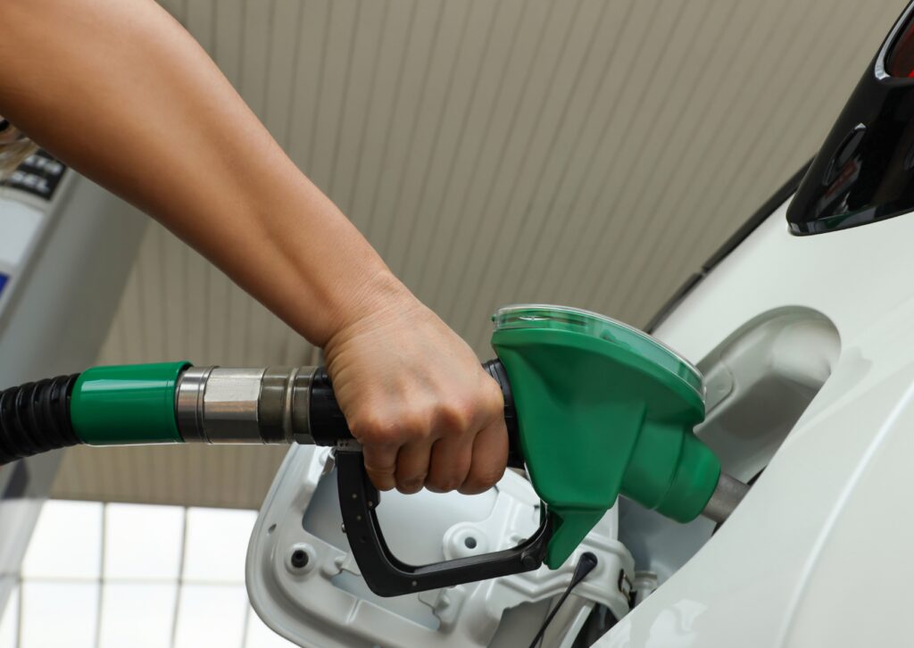 Driver refuels white car at gas station, hand gripping green fuel nozzle inserted into open gas cap area under canopy.