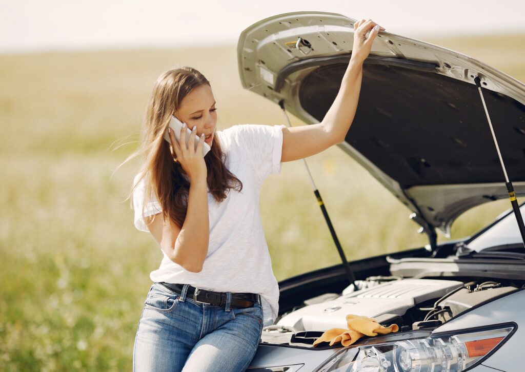 Woman calls for help beside car with hood up on roadside, leaning on vehicle while checking engine in open field.