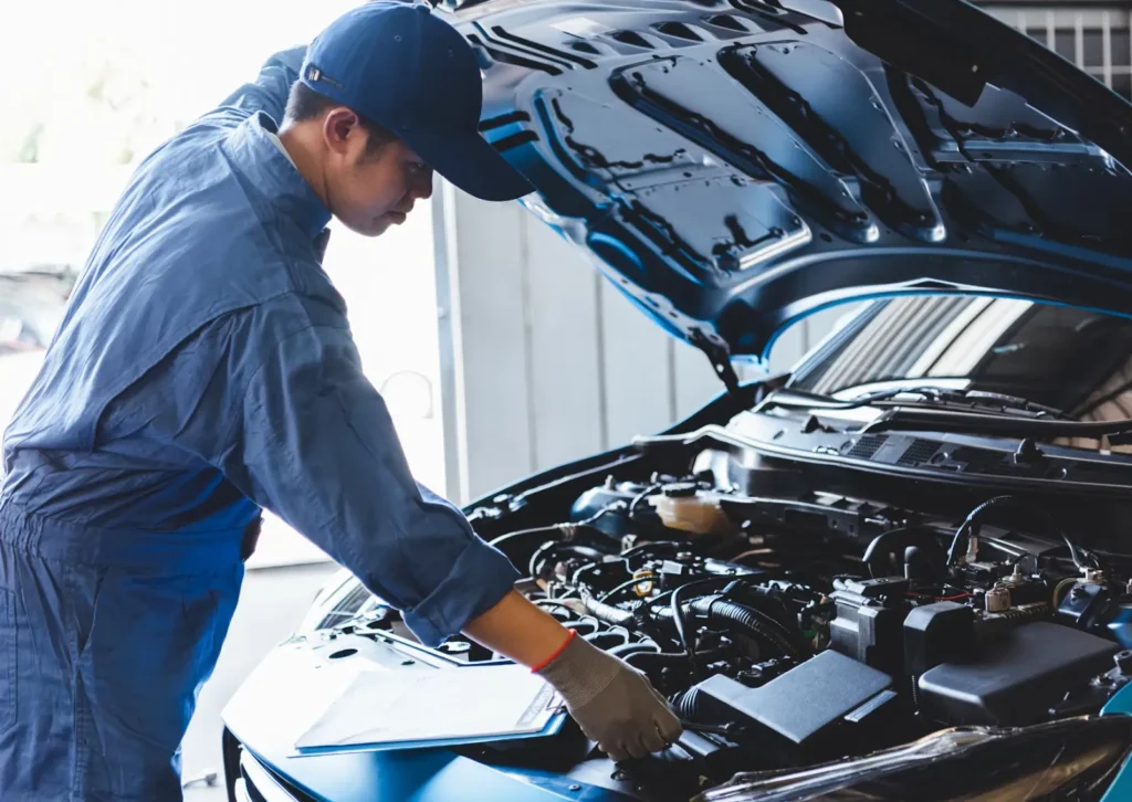 Auto mechanic in blue coveralls inspects engine bay under open hood, checklist clipboard on car fender.