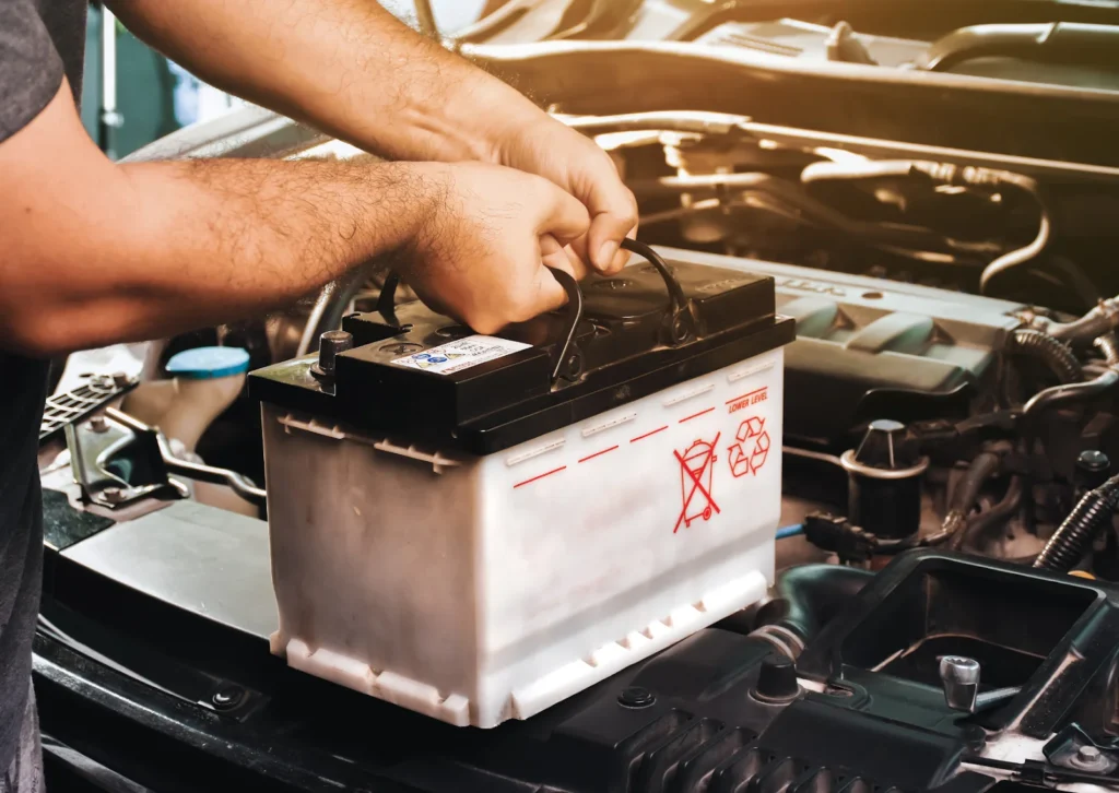 Hands lift a car battery from engine bay during replacement, with warning and recycle symbols visible.