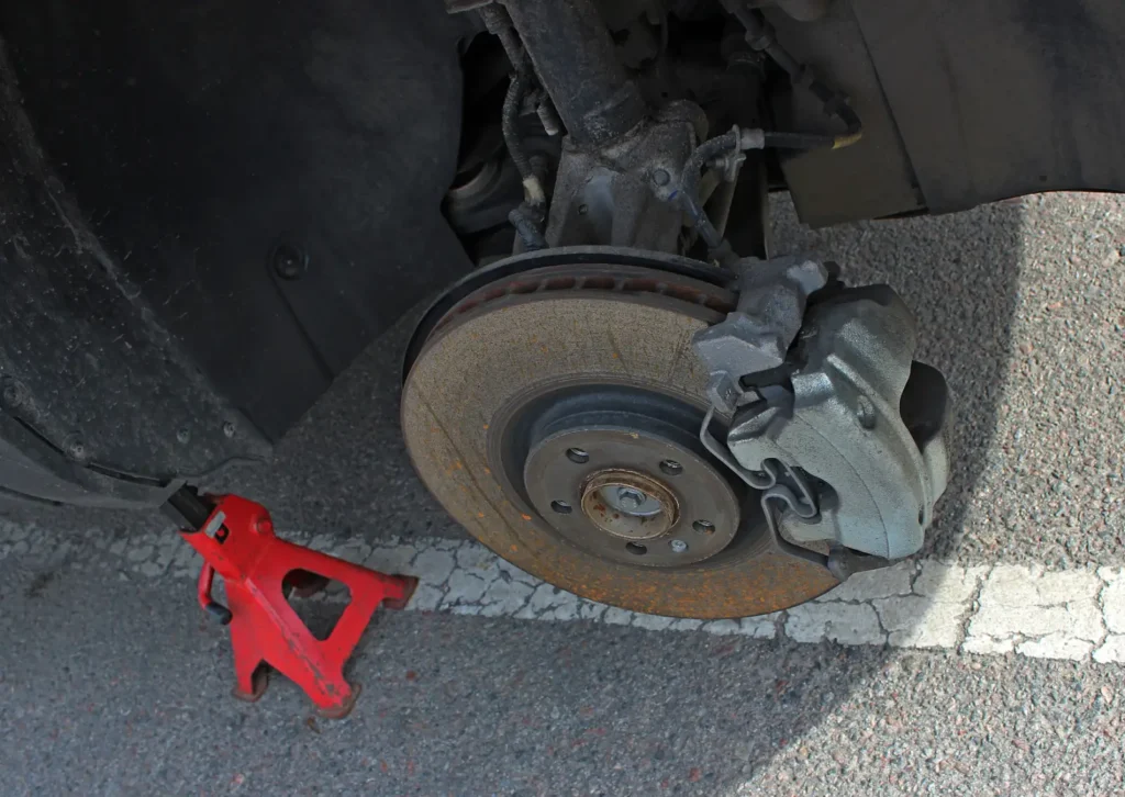 Car wheel removed showing brake rotor and caliper with light rust, supported by red jack stand on pavement.