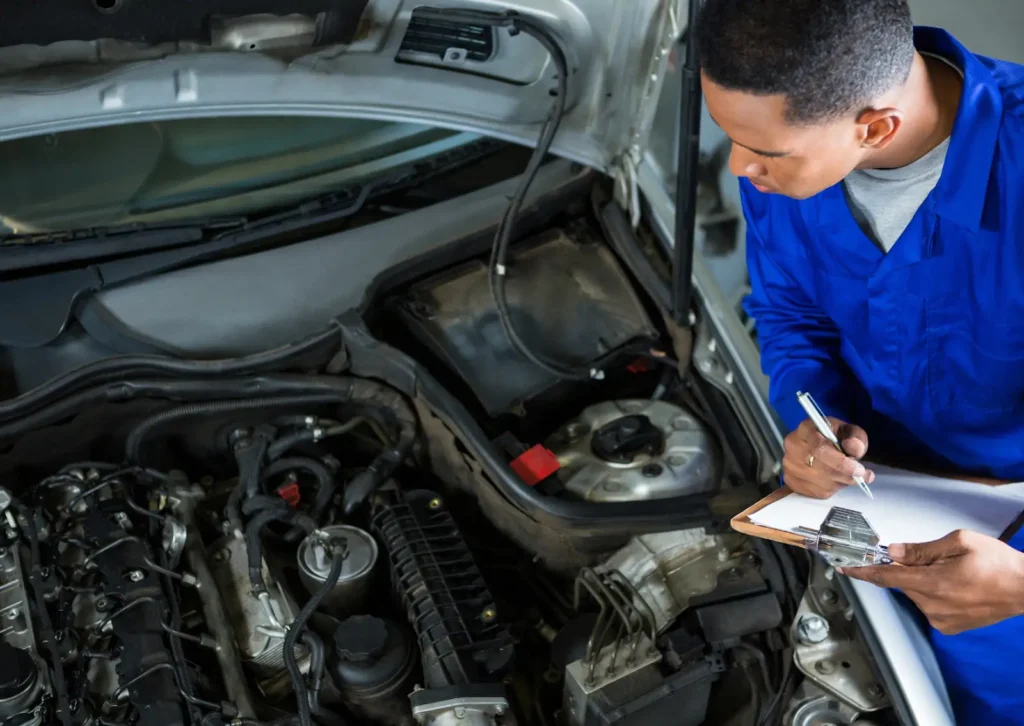 Mechanic in blue uniform inspects open car engine bay and writes notes on clipboard during routine service.