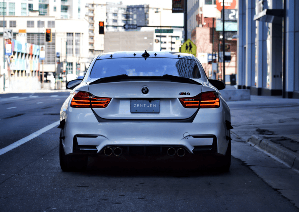 Rear view of a white BMW M4 with illuminated taillights parked on a quiet downtown street. 
