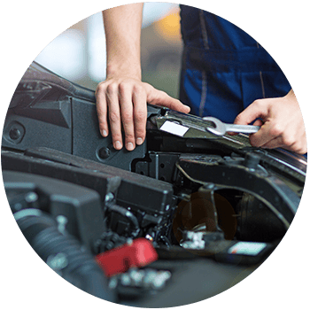 Circular photo of a mechanic in blue uniform using a wrench while working under a car’s open hood in a garage.