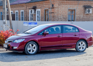 Maroon Honda Civic sedan with alloy wheels parked on sunlit street before brick building and grey fence.