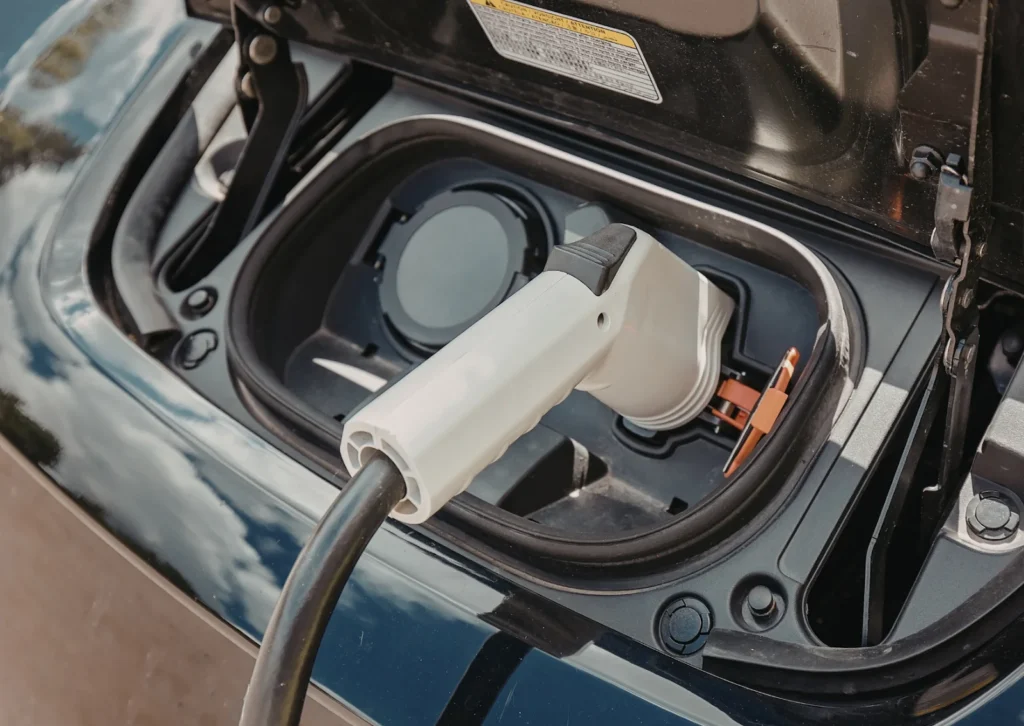 Close-up of electric vehicle charging connector plugged into port under open flap on dark car hood surface.
