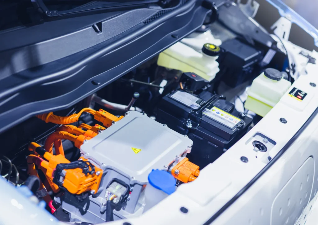 Electric vehicle engine bay with orange high-voltage cables, inverter unit, 12V battery, and coolant tanks.