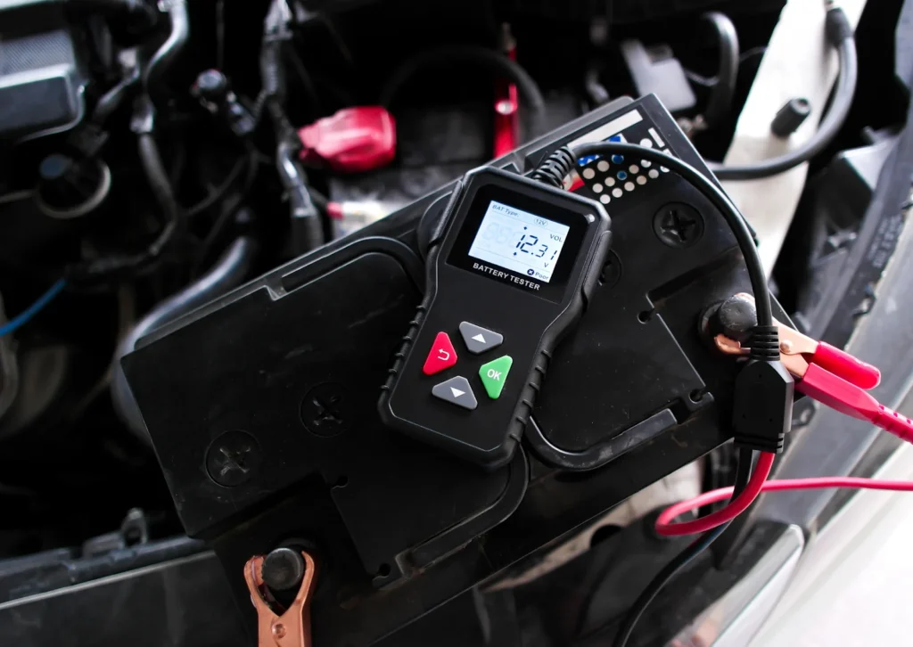 Battery tester clipped to car battery terminals shows 12.3 volts during under-hood electrical check.