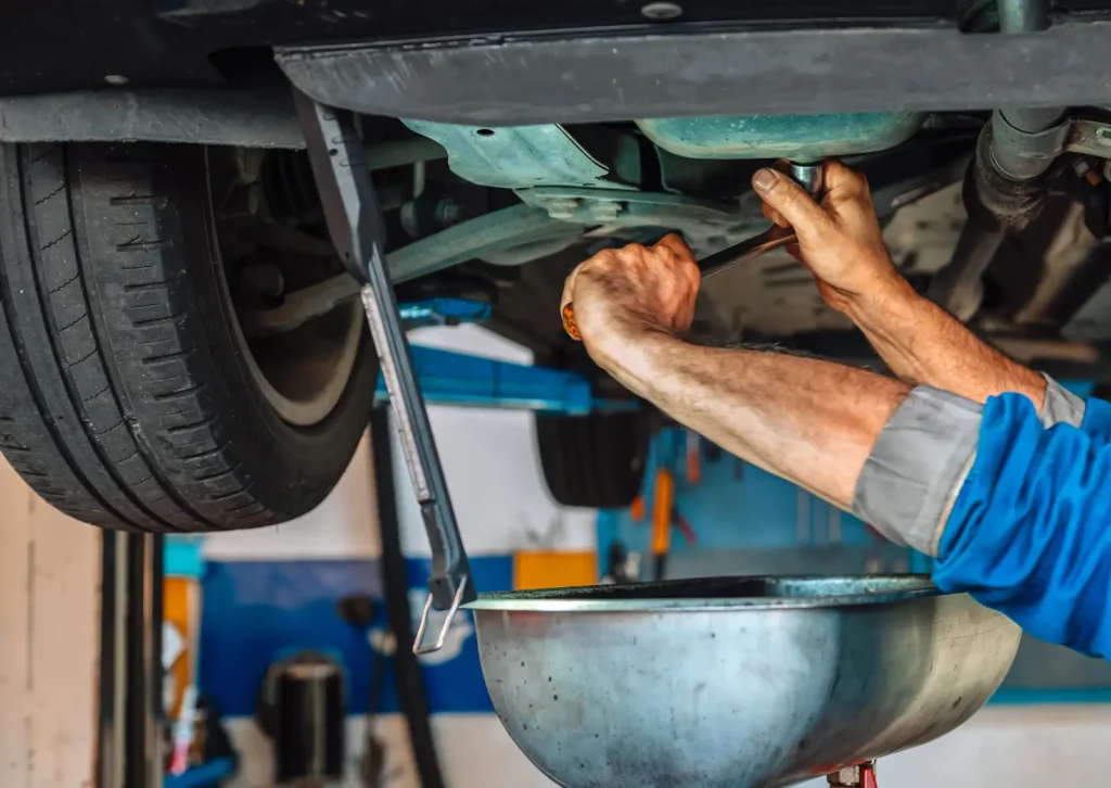 Mechanic loosens oil drain plug under lifted car, using wrench while drain pan below collects used oil.