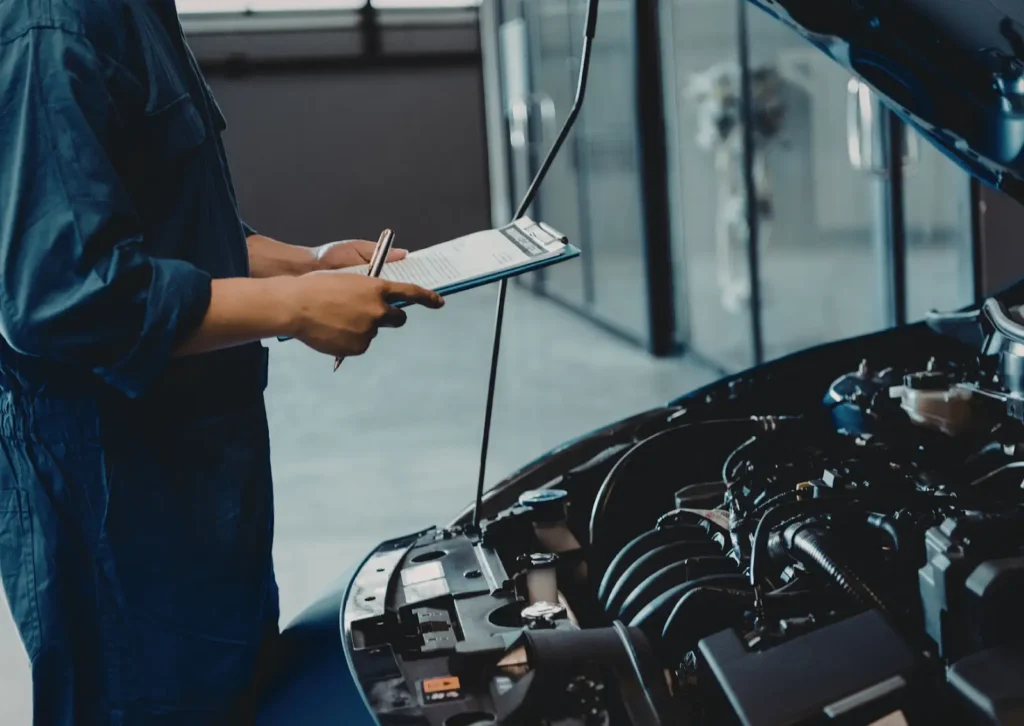 Mechanic in blue coveralls holds clipboard and pen while inspecting car engine bay in service workshop.
