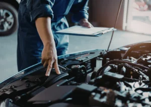 Auto mechanic in blue coveralls points at engine components while holding a clipboard in a busy garage.