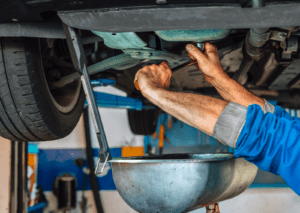 Mechanic loosens drain plug under lifted car, showing what causes oil leaks in car during service.