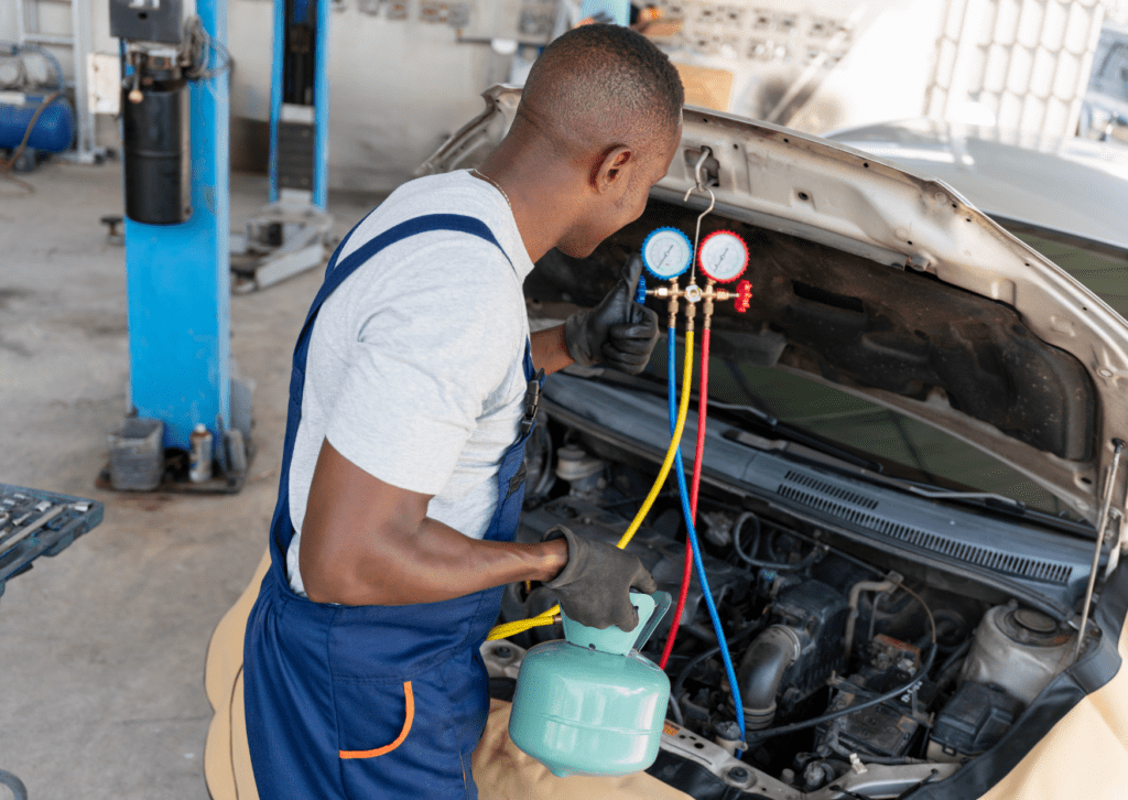 Mechanic servicing car AC system with manifold gauges and refrigerant tank under open hood 