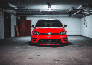 Front view of a red Volkswagen parked inside a dim concrete garage beneath exposed ceiling pipes. 